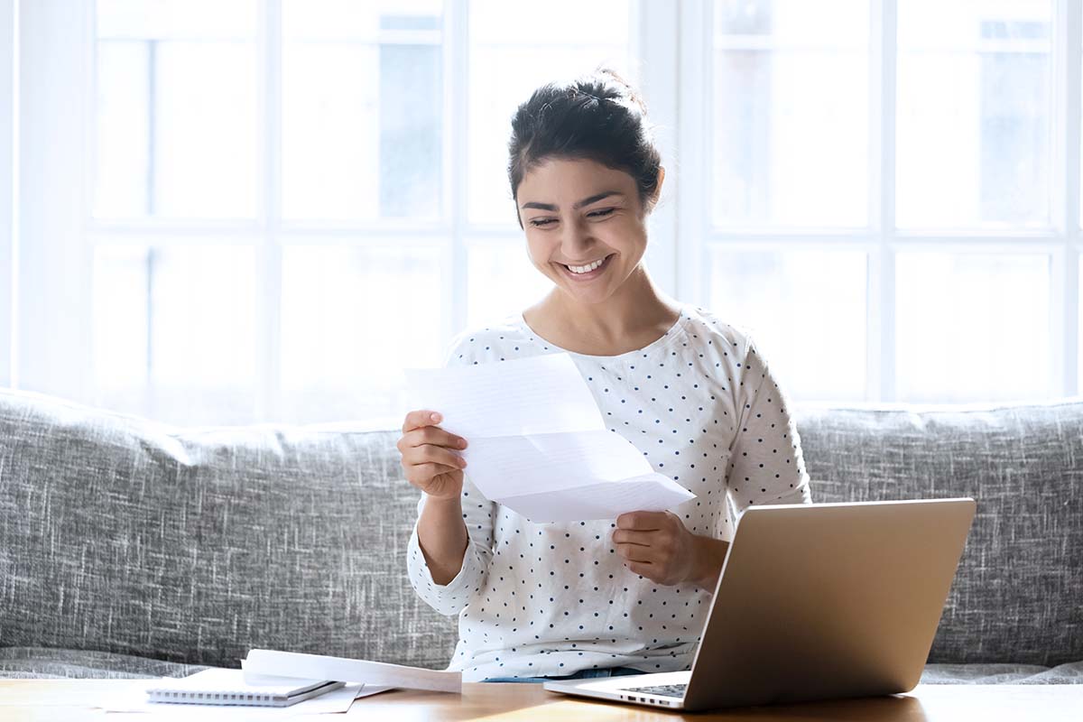 Young woman sitting on couch with laptop smiling at financial aid letter
