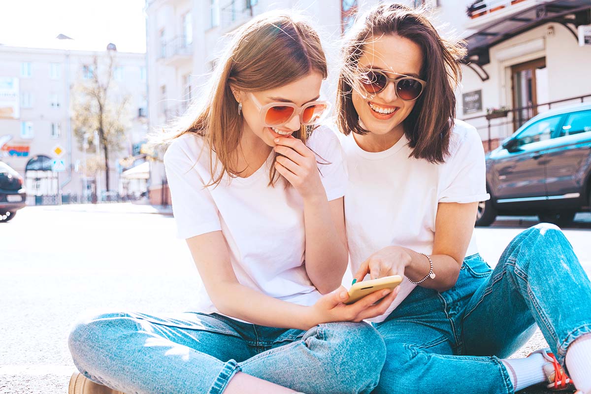 Two young women sitting on sidewalk looking at mobile phone